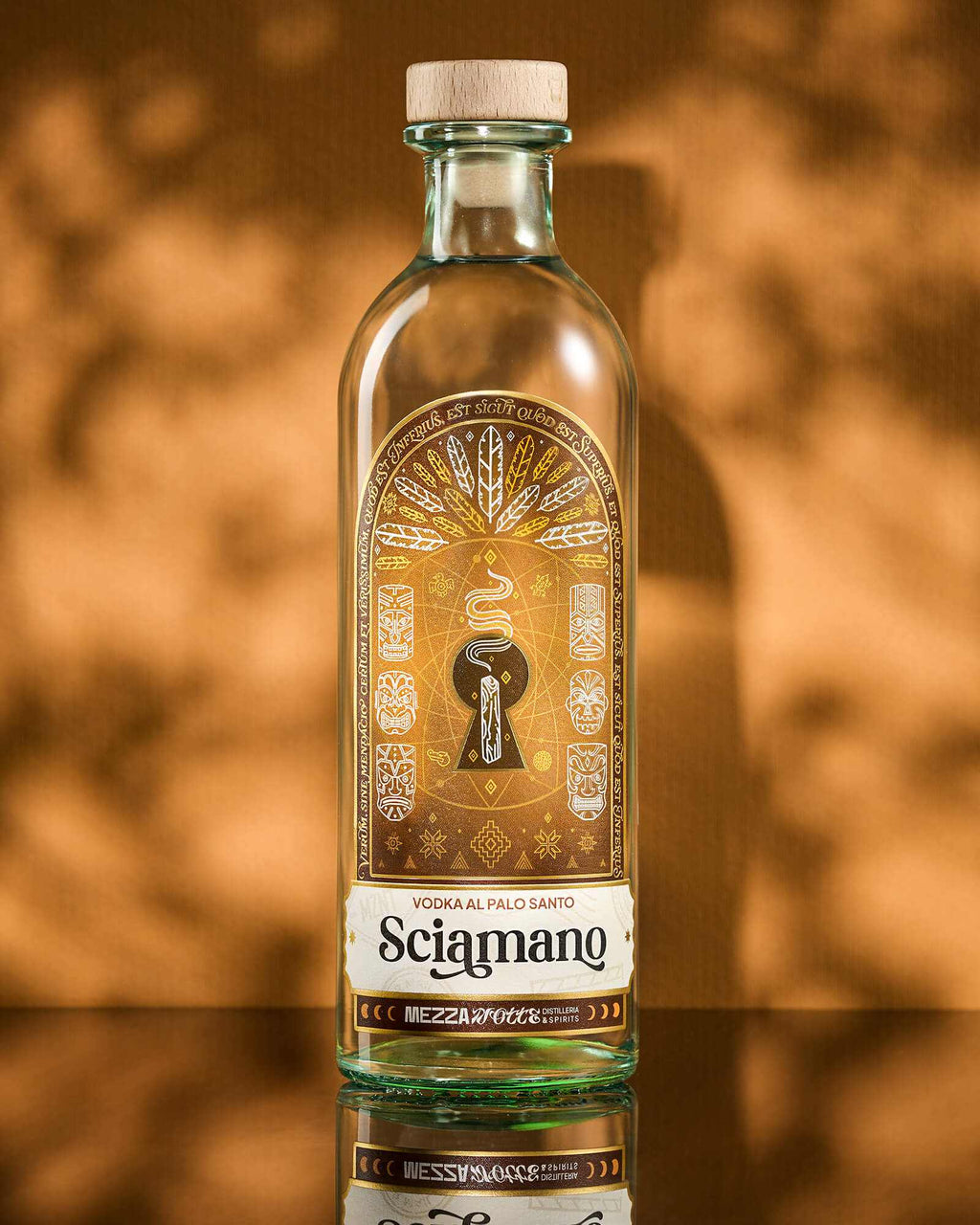 Bottiglia di Sciamano Vodka al Palo Santo Mezzanotte con etichetta dorata raffigurante maschere tribali e piume, su sfondo ambrato. Vodka aromatizzata artigianale italiana.